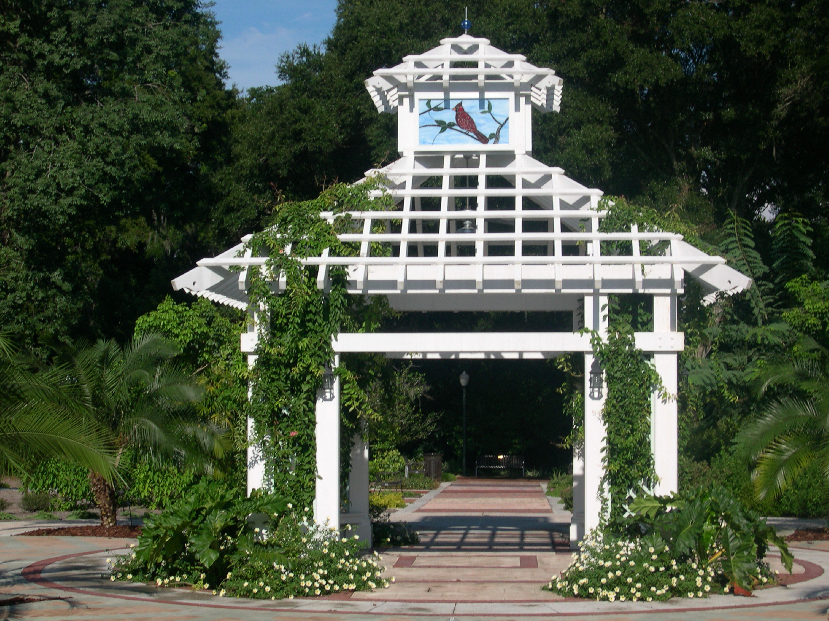 A white gazebo with a stained glass cardinal at the top in a courtyard surrounded by plants and flowers
