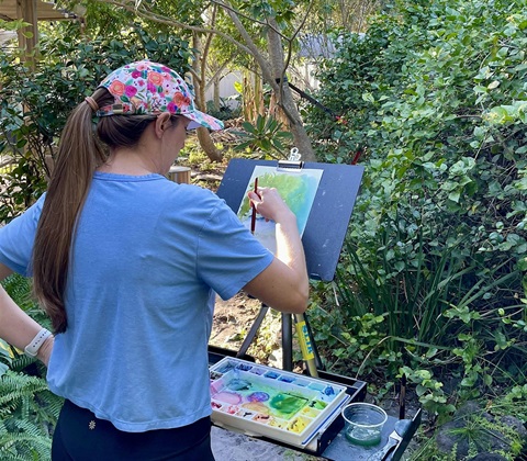 Person painting outside in a Garden. There are green trees around them. The person is wearing blue and has an art easel in front of them, with their painting on it and paints below it.